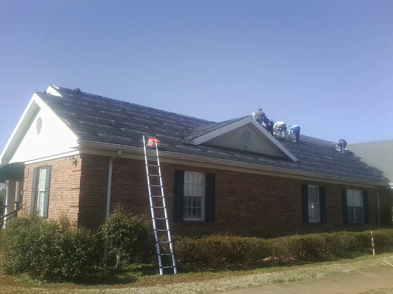 Crew tearing shingles off a brick ranch roof with a ladder set — Emergency Roof Repair work by our crew in Ponte Vedra